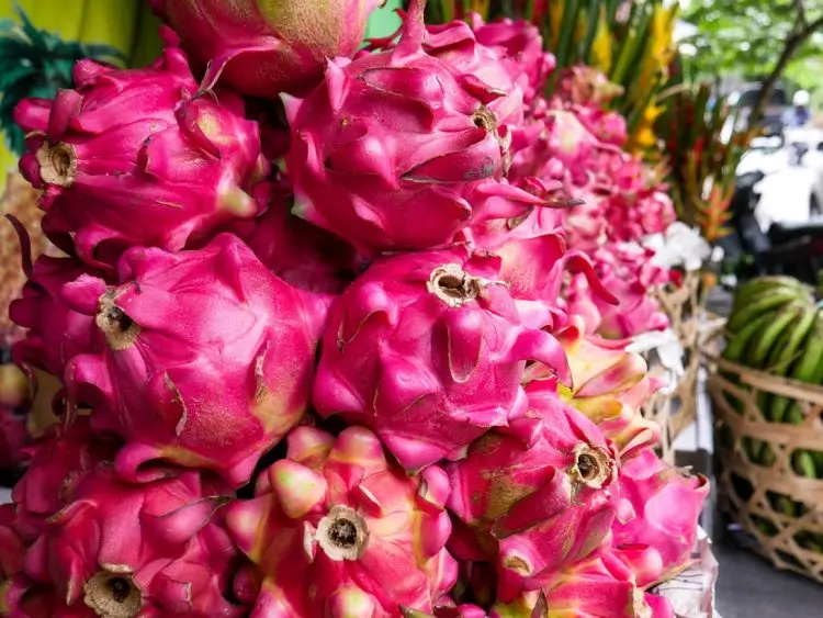 A close-up of a vibrant pile of pink dragon fruit with green-tipped scales at an outdoor market, with other tropical fruits and baskets visible in the blurred background.