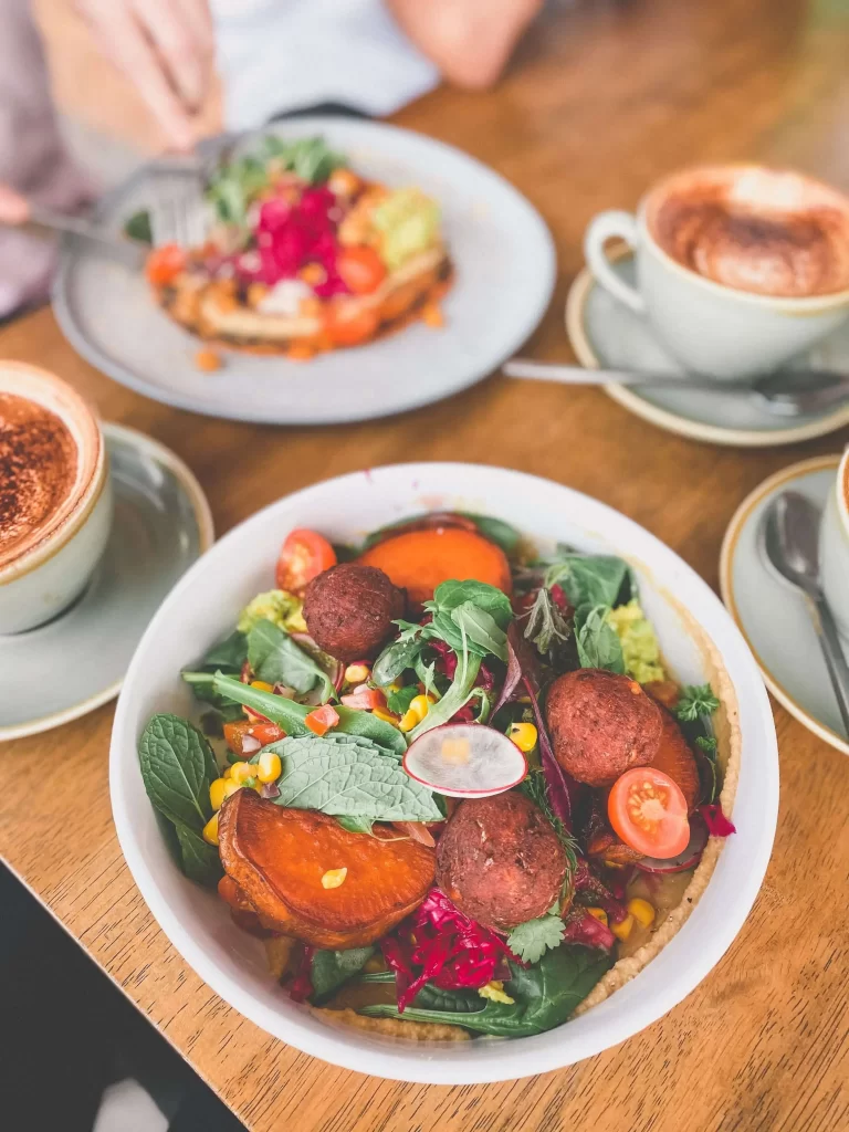 A colorful salad bowl with falafel, greens, tomatoes, sweet potato, corn, and beet is beautifully presented alongside two cups of cappuccino and a plate of toast on a wooden table at Mocha Central Coast Cafe.