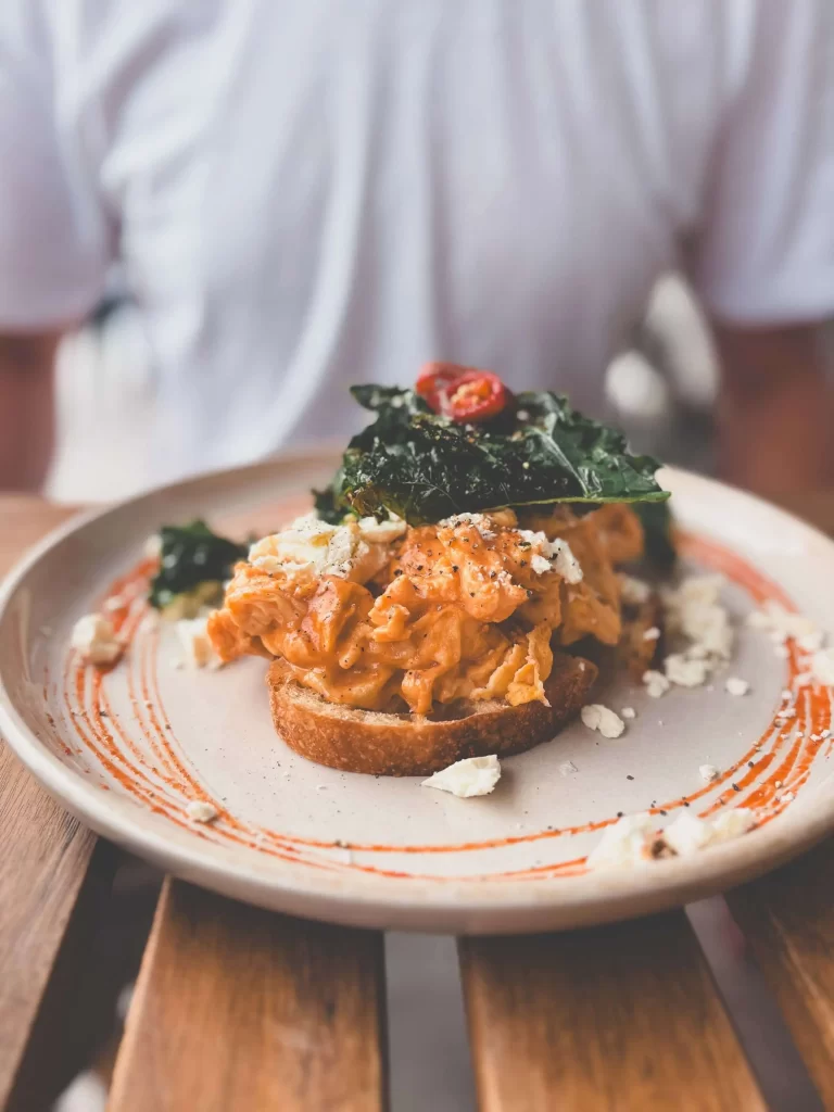 A plate with toast topped with scrambled eggs, crumbled cheese, leafy greens, and a cherry tomato, garnished with sauce— a classic offering found at many Central Coast cafes. A person in a white shirt sits behind the dish at a wooden table, perhaps savoring the ambiance of the morning coffee society.