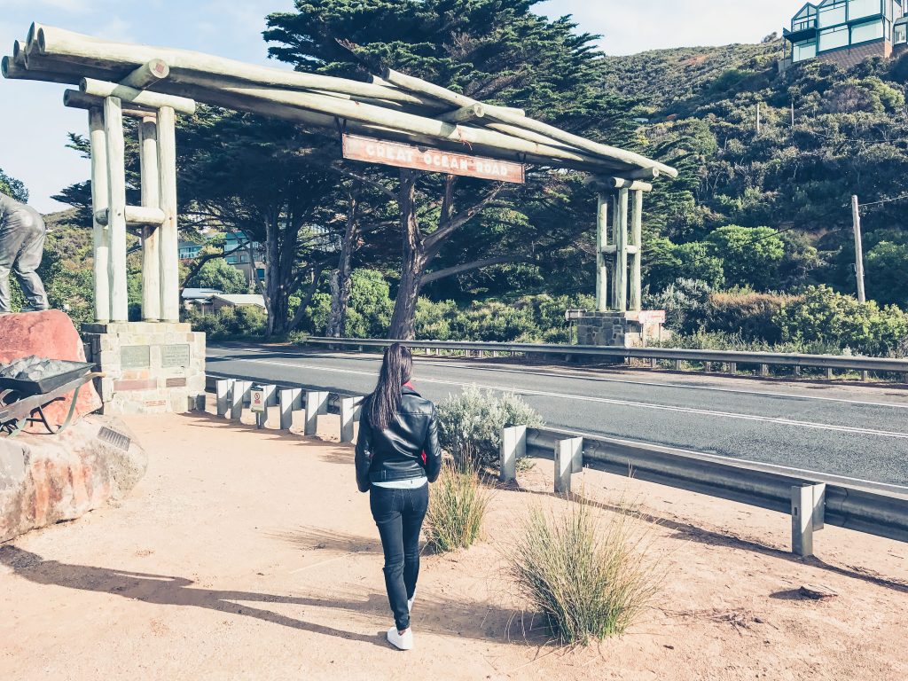 A person with long dark hair, wearing a black jacket and jeans, walks toward the large wooden Great Ocean Road archway sign beside a road surrounded by greenery and trees—a perfect beginning to any great ocean road itinerary.