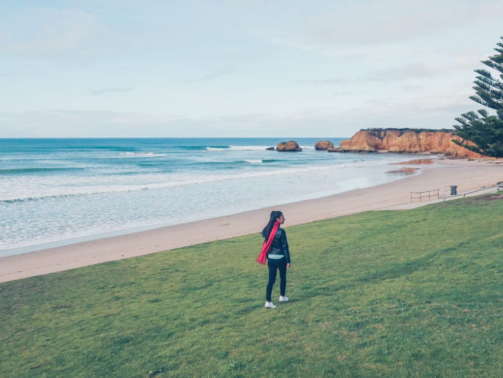 A person with a red bag stands on a grassy area, gazing out at a sandy beach and the calm ocean—an iconic stop along any Torquay Great Ocean Road itinerary. Rocky cliffs and a few trees are visible in the distance under a partly cloudy sky.