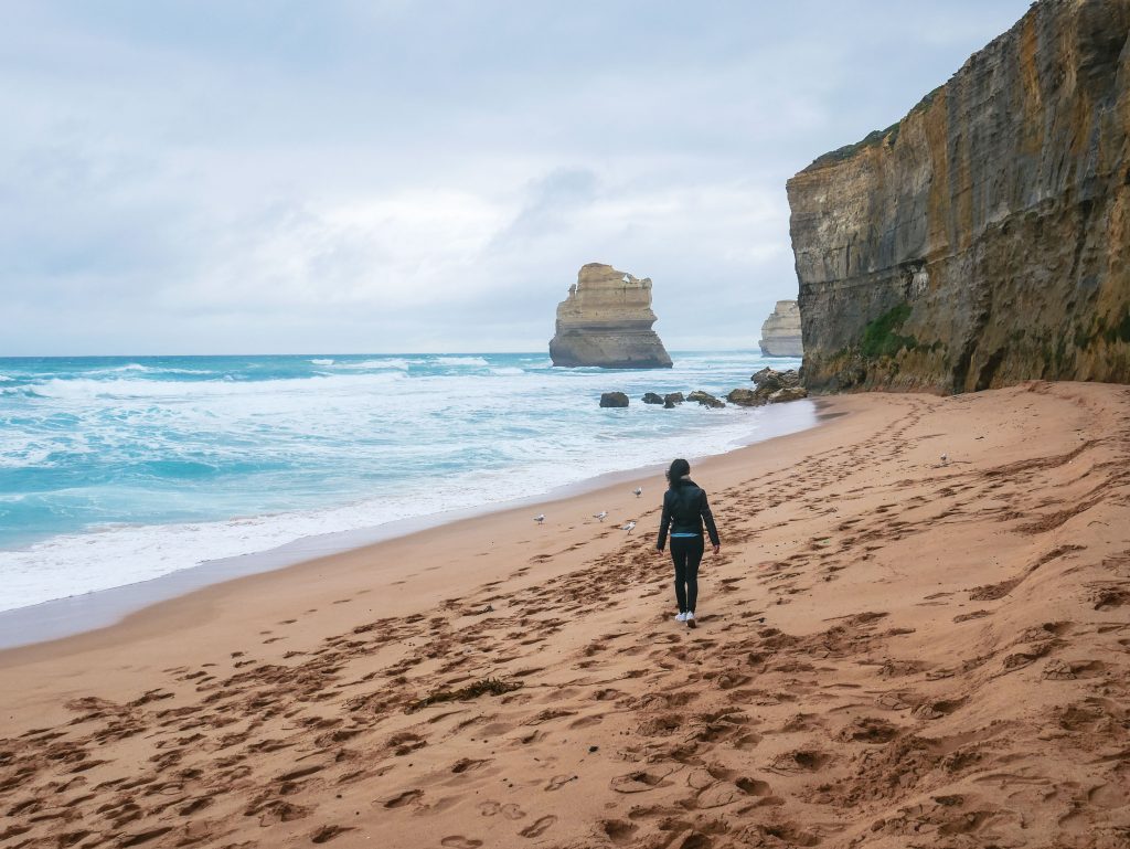 A person walks alone on a sandy beach near rocky cliffs with ocean waves crashing, reminiscent of the dramatic landscape at Gibson Steps. Footprints trail behind them and a large rock formation stands in the water under a cloudy sky.