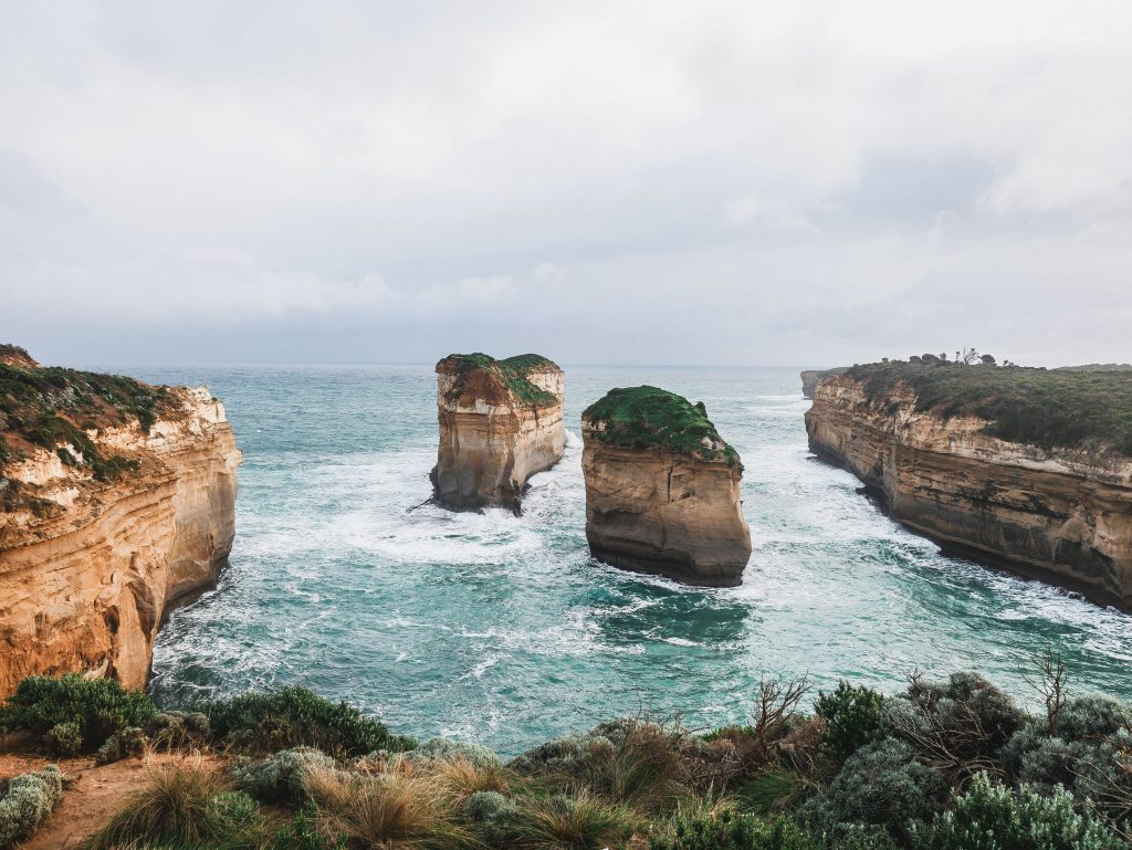 Two large rock formations stand in turquoise ocean water near the rugged coastline of Loch Ard Gorge, with waves crashing around them and greenery in the foreground under a cloudy sky.