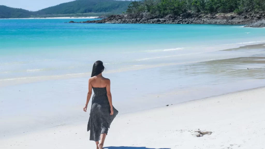 A woman in a black dress walks along the white sands of Whitehaven Beach beside clear turquoise water, with green hills and trees in the background under a bright sky.