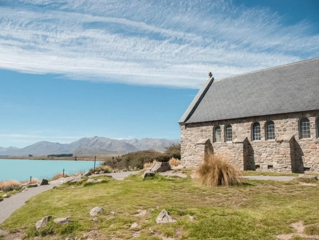 A stone church with arched windows sits by the turquoise waters of Lake Tekapo, with mountains and a blue, cloud-streaked sky in the background. Grassy ground and a dirt path lead toward the water.