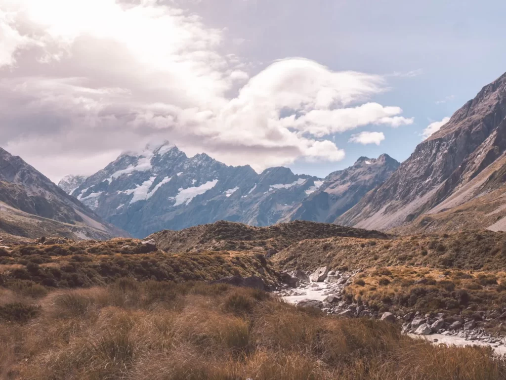 Snow-capped mountains, including the majestic Mount Cook, rise under a partly cloudy sky, with grassy hills and a rocky stream winding through the foreground.