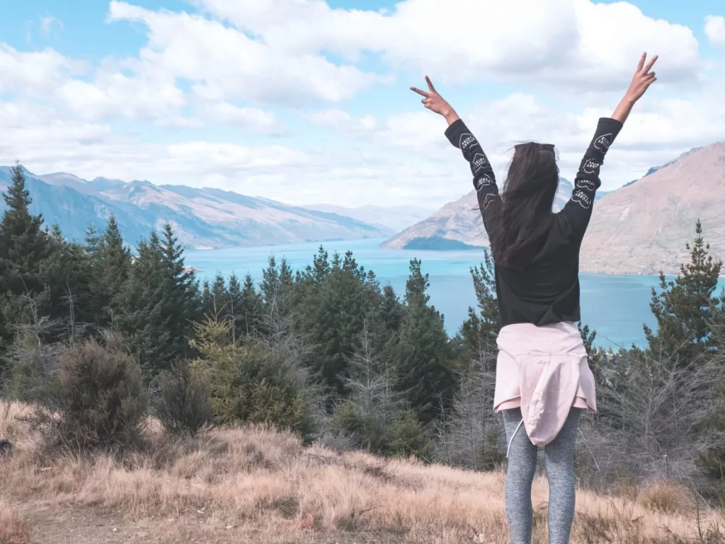 A person stands on Queenstown Hill with arms raised in a peace sign, overlooking a lake surrounded by trees and mountains under a cloudy sky.