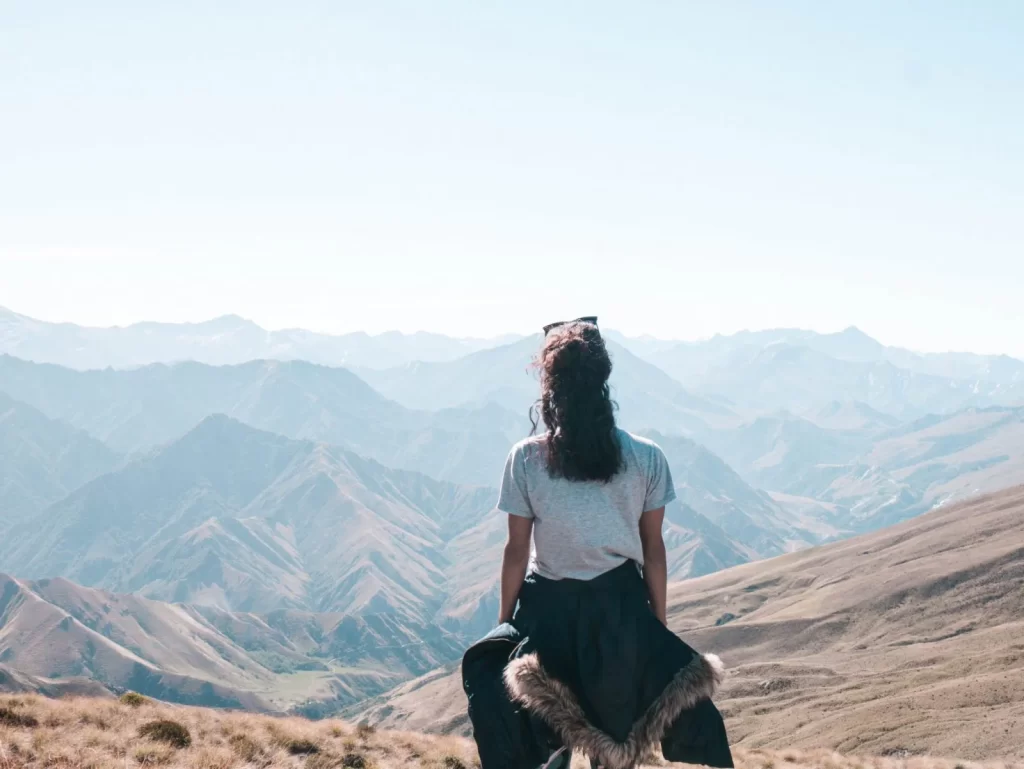 A person with long hair sits on a hilltop near the Ben Lomond Summit, facing distant, hazy mountains under a clear sky, wearing a gray shirt and a dark jacket tied around their waist. Grassy slopes and ridges stretch out below.