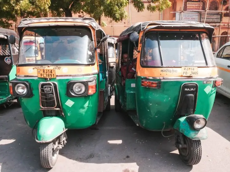 Two green and yellow auto rickshaws, also known as tuk tuks, are parked side by side on a city street under sunlight, with buildings and trees visible in the background—a typical scene when exploring the best things to do in Jaipur.