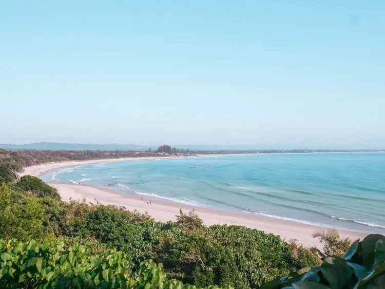 A wide sandy beach curves along calm blue water, bordered by green vegetation and distant trees under a clear, bright sky—capturing the tranquil beauty of Main Beach Byron Bay.