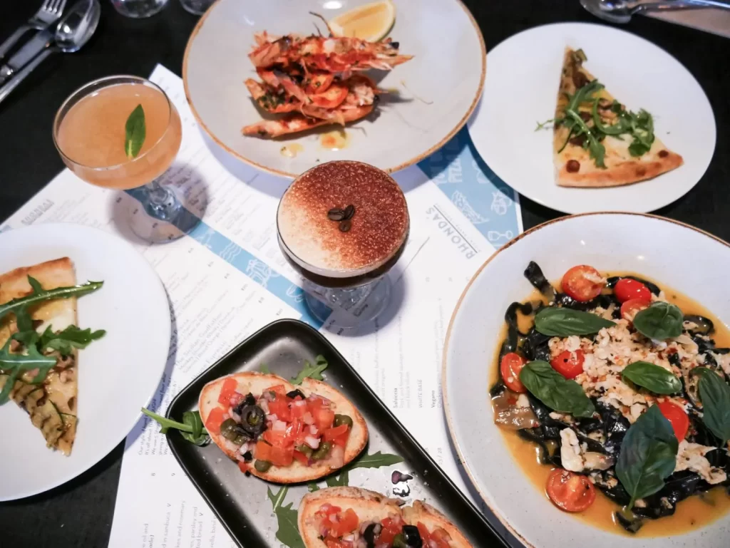 An overhead view of a table at Rhonda's Bar Terrigal restaurant, featuring shrimp, bruschetta, pasta, two slices of pizza, a cocktail, and a dessert—all served on white plates and set atop a menu.