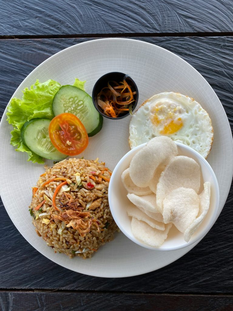 A plate of Indonesian food featuring a mound of fried rice topped with crispy shallots, served with a sunny side up egg, a bowl of prawn crackers, and a side of fresh cucumber, tomato, and lettuce. A small dish of pickled vegetables adds color and texture to the meal. The dish is neatly arranged on a white plate over a dark wooden table.