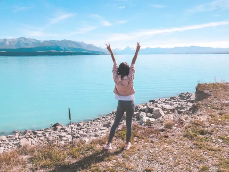 A person stands on a rocky shore of Lake Pukaki with arms raised, facing the calm blue lake and distant mountains under a clear sky.