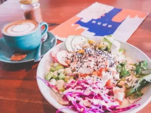 A bowl of colorful salad with shredded cabbage, radish, edamame, cucumber, avocado, and sesame seeds sits next to a cup of cappuccino on a wooden table at Canggu cafe Nude, with a menu partially visible in the background.