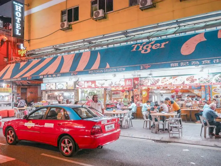 A busy street scene at night with a red taxi parked in front of Restoran Dragon View, where people are dining at outdoor tables under a blue Tiger-branded awning—a vibrant snapshot that perfectly captures the lively atmosphere recommended on any Kuala Lumpur itinerary.