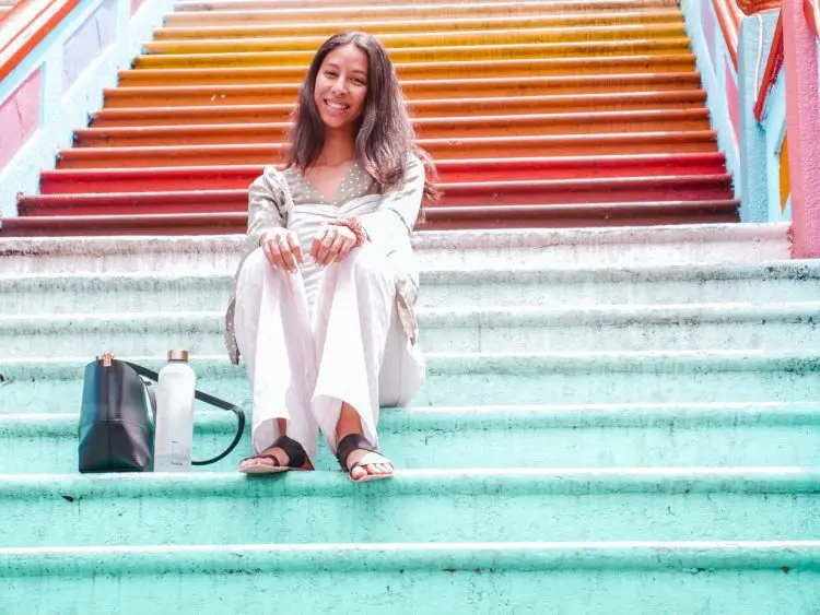 A woman sits smiling on colorful, rainbow-painted stairs, reminiscent of the vibrant Batu Caves often included in a Kuala Lumpur itinerary. She wears white pants and a light jacket, with a black bag and water bottle beside her. Bright sunlight illuminates the scene.