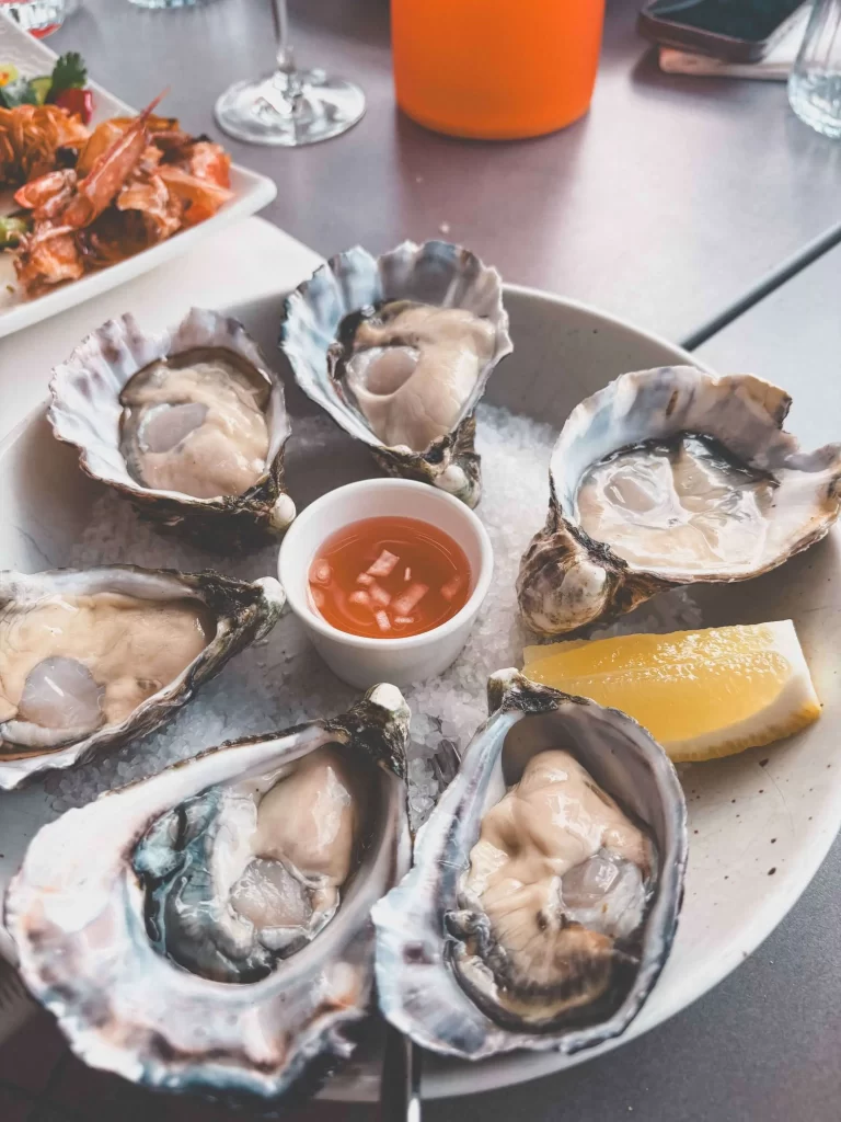 A plate with six raw oysters on the half shell, a lemon wedge, and a small cup of red sauce, set on a bed of ice at Swells Terrigal restaurant. Shrimp and drinks are visible in the background.