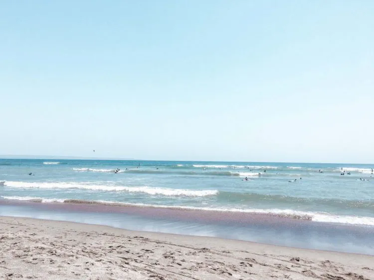 A sandy beach with gentle waves rolling in under a clear blue sky; several people are visible in the water, possibly surfing or swimming—an ideal stop on any Bali itinerary.