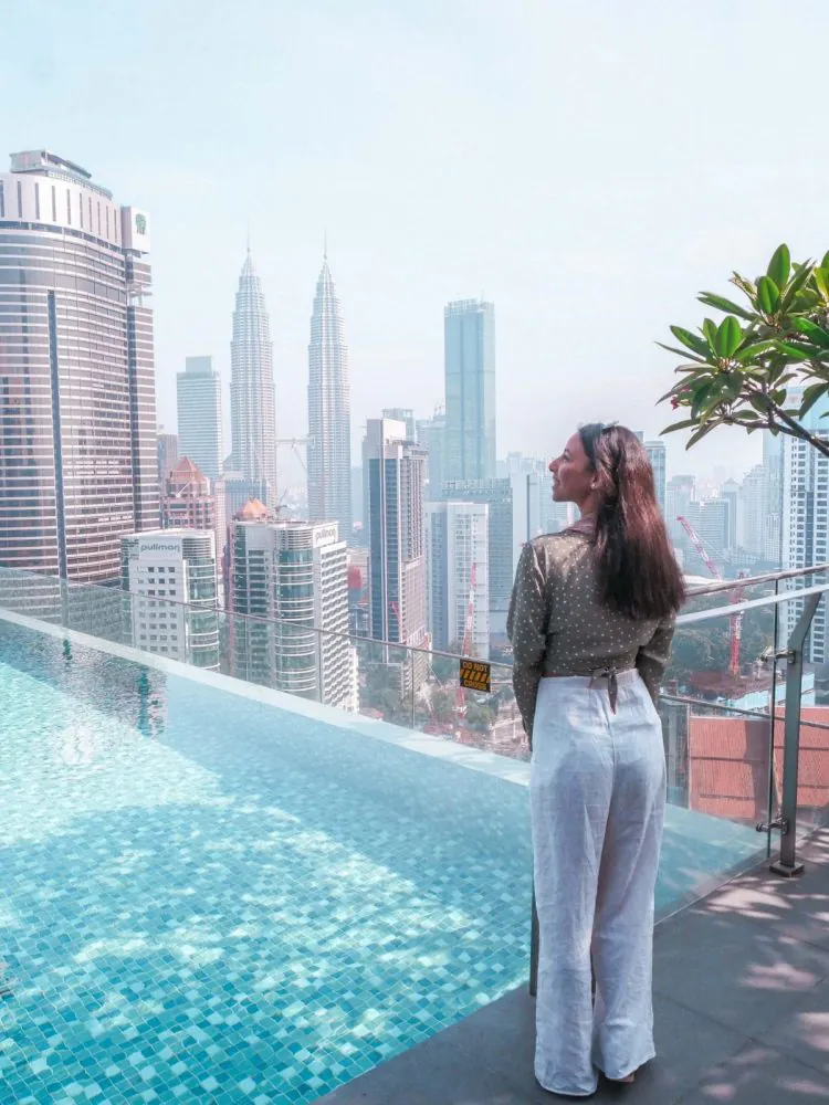 A woman stands by a rooftop pool, looking out at tall city buildings and the Petronas Towers in Kuala Lumpur on a hazy day—a perfect moment to begin her Kuala Lumpur itinerary.