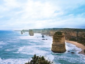 Rock formations known as the 12 Apostles rise from the blue ocean near a rugged coastline with sandy beaches and green shrubs under a cloudy sky.