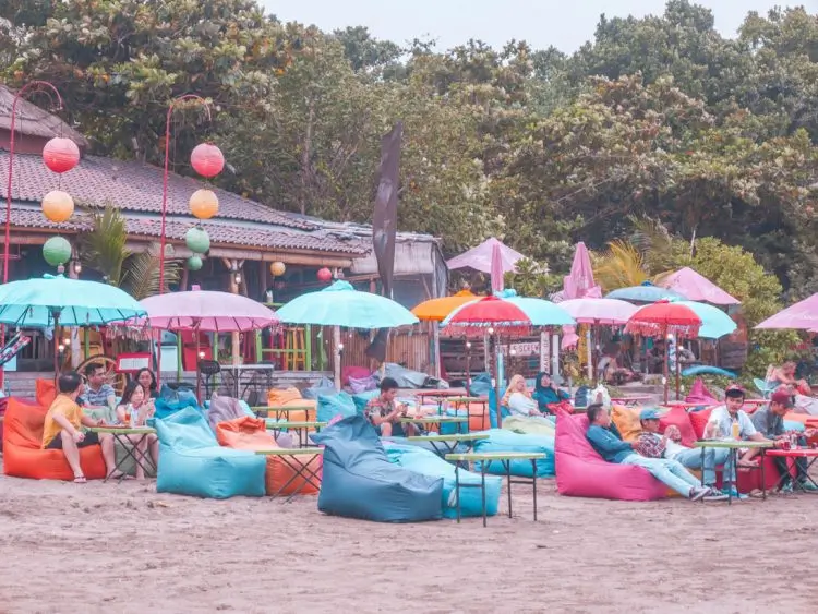 La Plancha - Colorful bean bags and tables with umbrellas are set up on a sandy Seminyak beach, perfect for relaxing and socializing. Trees and a vibrant building decorated with lanterns make this a must-see spot on any Bali itinerary.