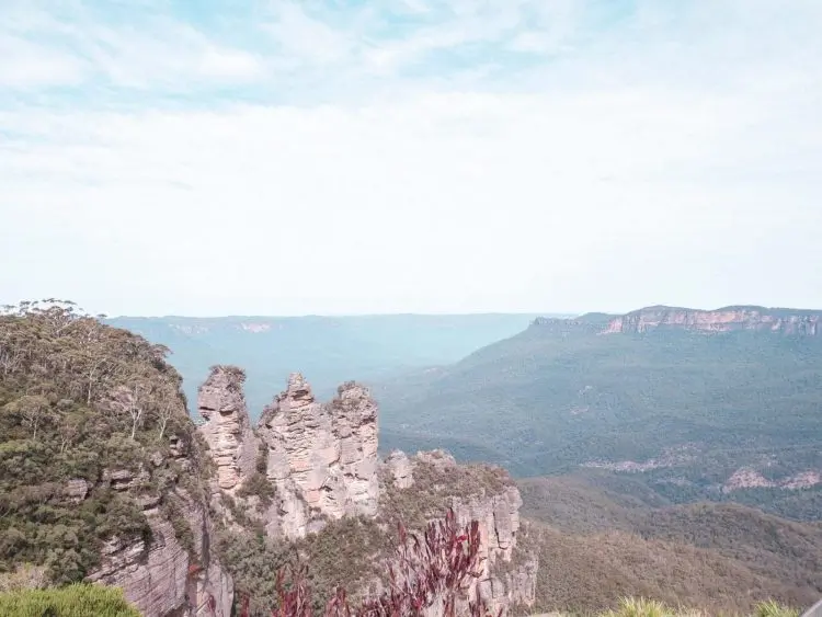 The image shows the iconic Three Sisters rock formation in the Blue Mountains, Australia, surrounded by lush green forest and distant blue-hued mountain ridges under a bright, cloudy sky.
