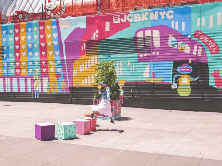 A person jumps over colorful cubes in front of a vibrant mural featuring hearts, stripes, and playful cartoon characters, with JCBKNYC written on the wall—an eye-catching stop perfect for any New York itinerary exploring the artistic side of the Financial District.