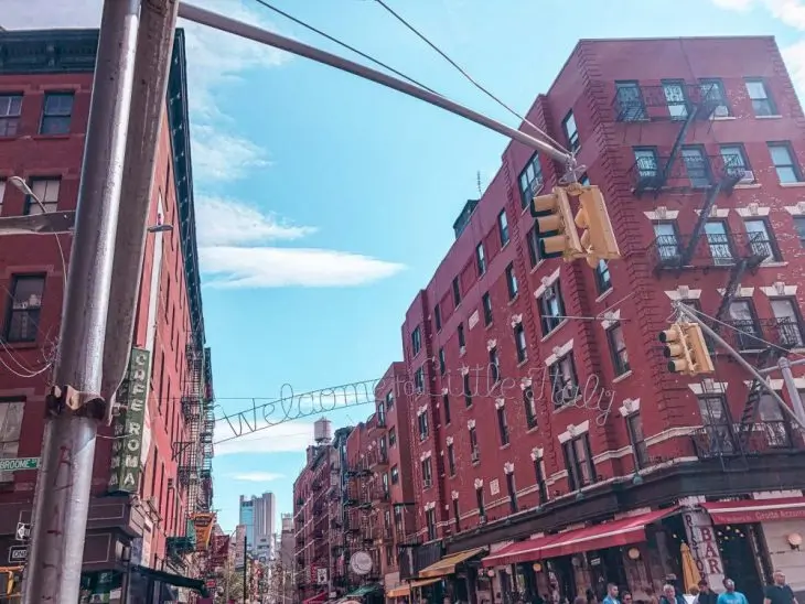 A city street with red brick buildings, traffic lights, restaurants, and shops under a blue sky evokes the vibrant charm of Little Italy—a must-see on any New York itinerary. Pedestrians walk along the sidewalk, adding to the urban, lively atmosphere.
