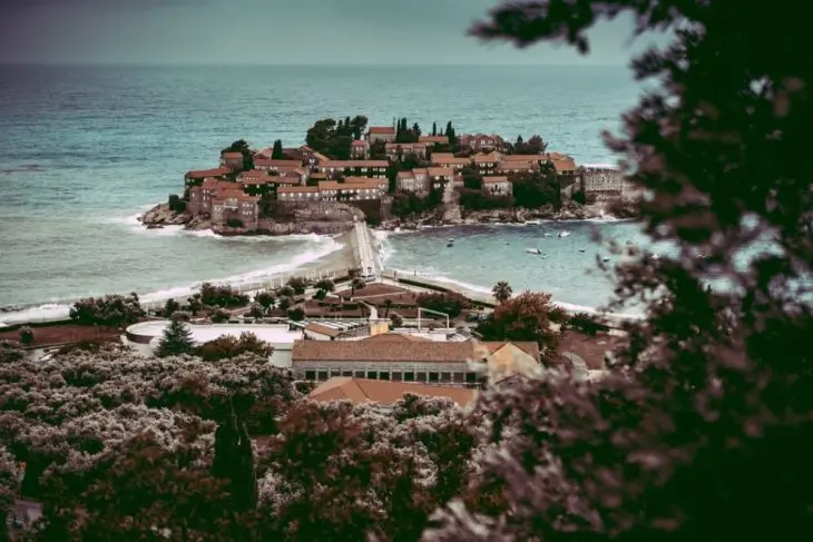 An island with red-roofed buildings is connected to the mainland by a narrow causeway, surrounded by the blue sea and greenery under a cloudy sky—a stunning highlight on any Montenegro itinerary.