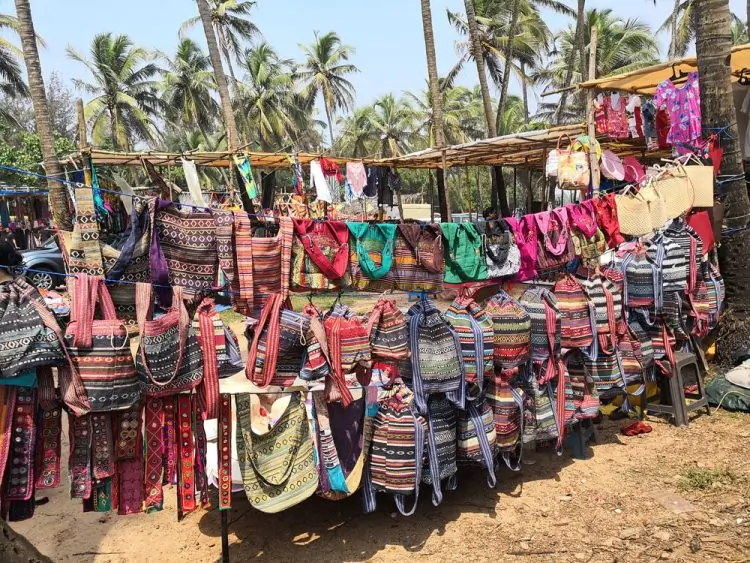 A market stall displays colorful, patterned bags and purses hanging in rows, with palm trees and a blue sky in the background—an inviting scene to add to your Goa itinerary.