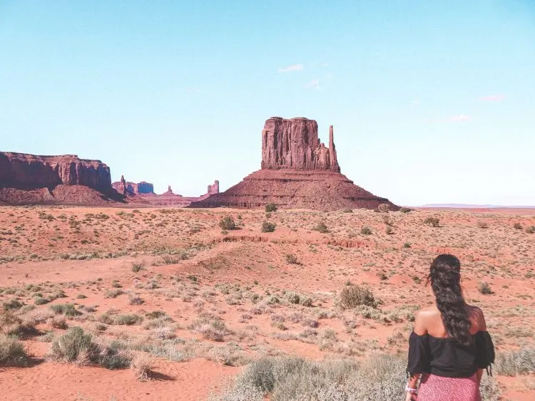 A woman with long dark hair stands in the desert, gazing at a large, flat-topped butte in Monument Valley under a clear blue sky.