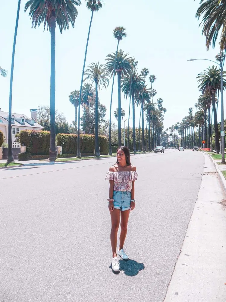 A young woman in a floral top and denim shorts stands on a sunny, empty street lined with tall palm trees and houses in Beverly Hills.