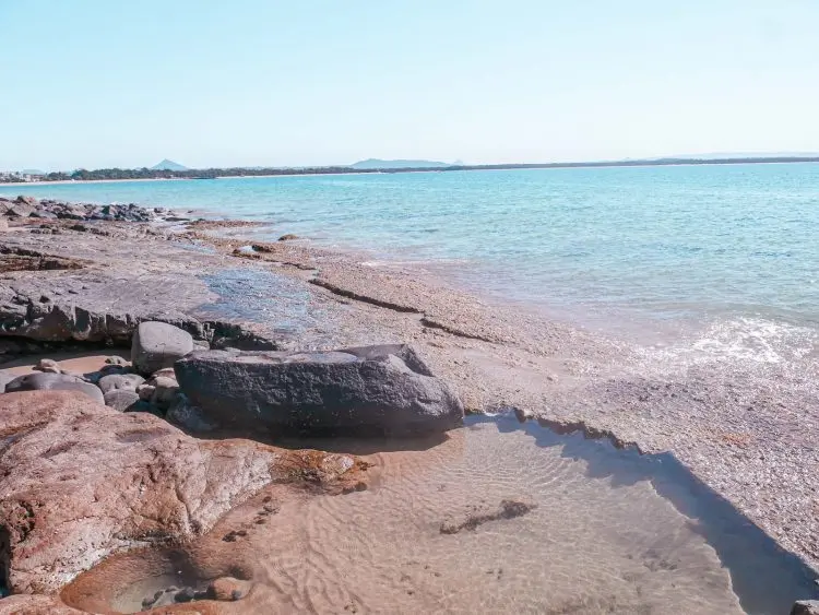 Rocky shoreline with shallow pools and gentle waves, clear blue water stretching to the horizon under a bright sky, distant land visible across the water—an iconic coastal scene reminiscent of Noosa National Park.