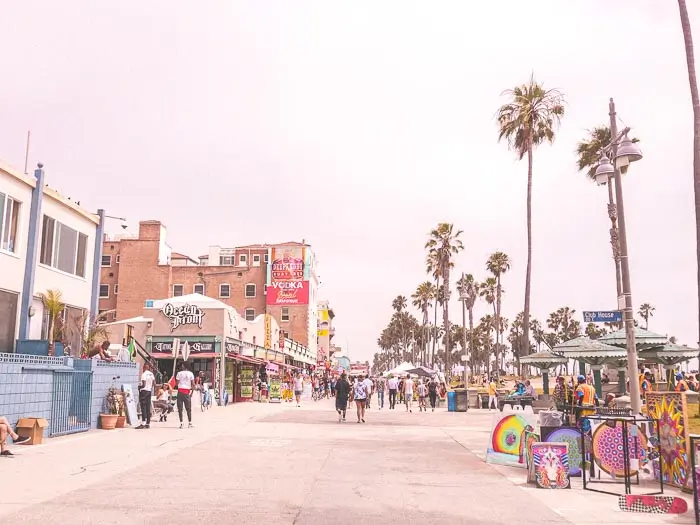 People stroll along a wide pedestrian path lined with shops, palm trees, and colorful street art on a bright, sunny day at Venice Beach, California.