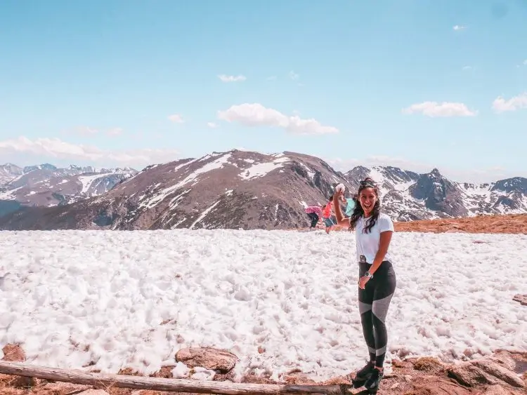 A woman in athletic wear stands smiling on snow with the majestic Rocky Mountains in the background under a clear blue sky. Another person is in the distance, holding their arms out, surrounded by the scenic landscape.