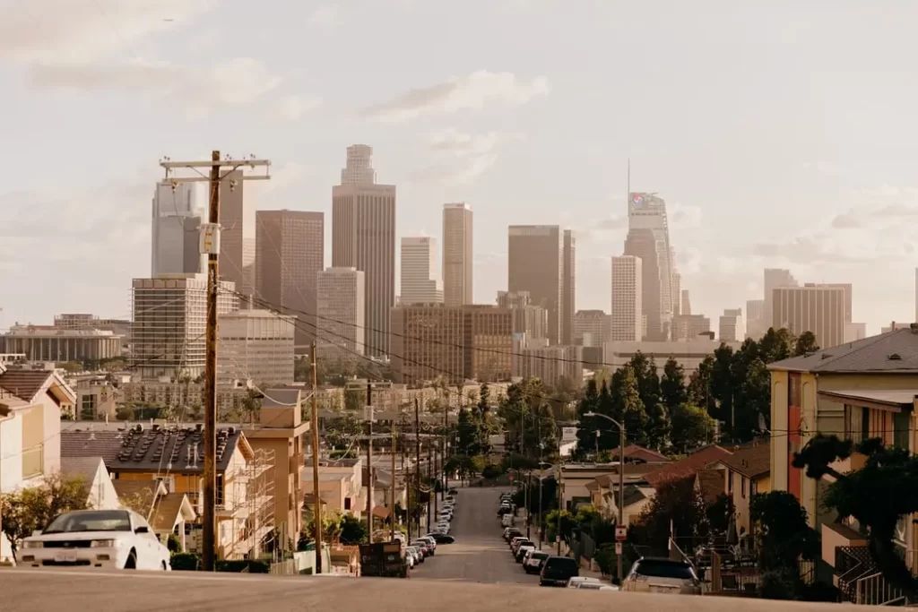 A city street lined with houses leads toward a downtown Los Angeles skyline filled with tall buildings under a partly cloudy sky. Power lines cross the scene, and trees are scattered among the homes.