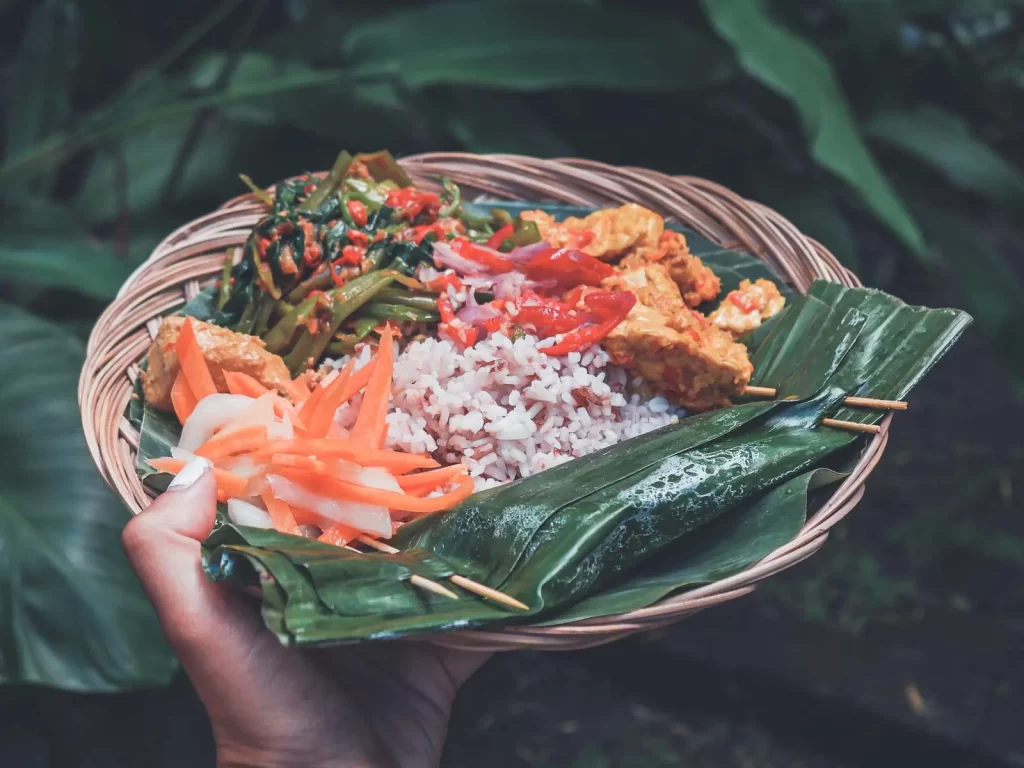 A hand holds a woven plate with rice, vegetables, tempeh, pickled carrots, and greens, all partially wrapped in banana leaves, against a background of green foliage—offering a glimpse of what you might create in a cooking class, one of the best things to do in Bali.