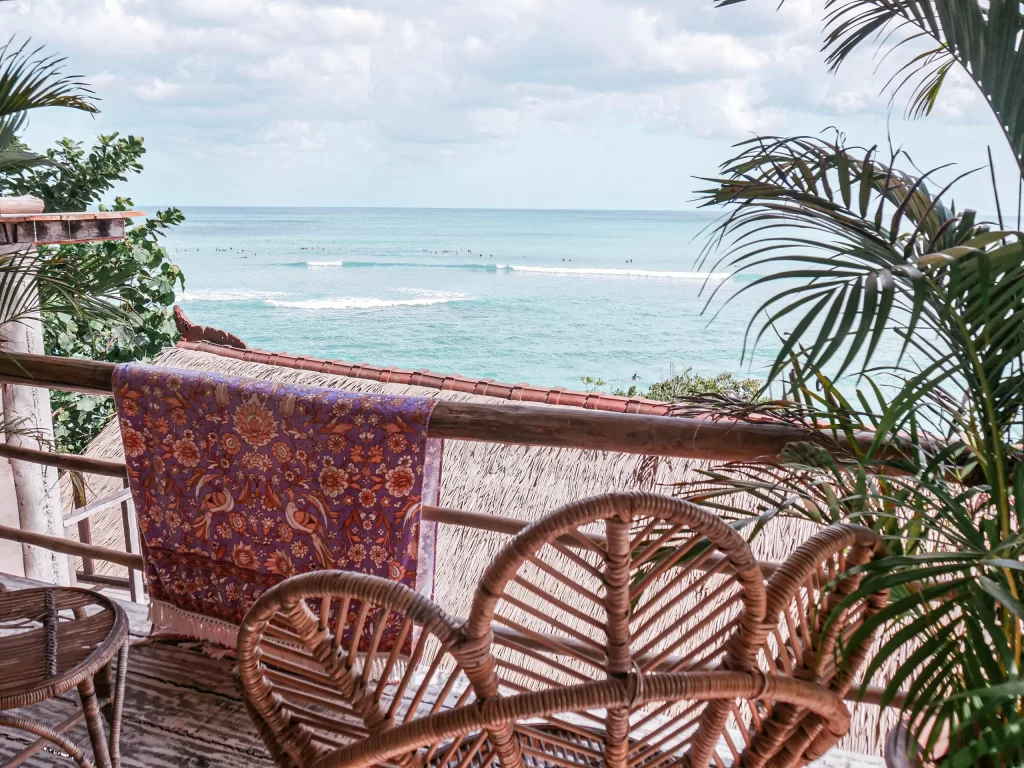 Wicker chairs and a table on a balcony with a purple patterned towel draped over the railing, overlooking the ocean with gentle waves and palm leaves framing the scene—a tranquil spot inspired by the serene beauty of Uluwatu Beaches.