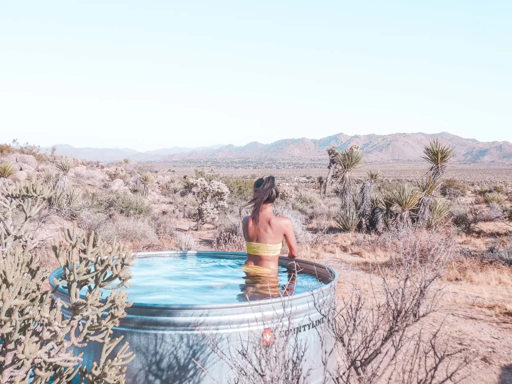 A person in a yellow swimsuit sits in a metal tub filled with water, surrounded by desert plants like joshua trees and distant mountains under a clear sky.