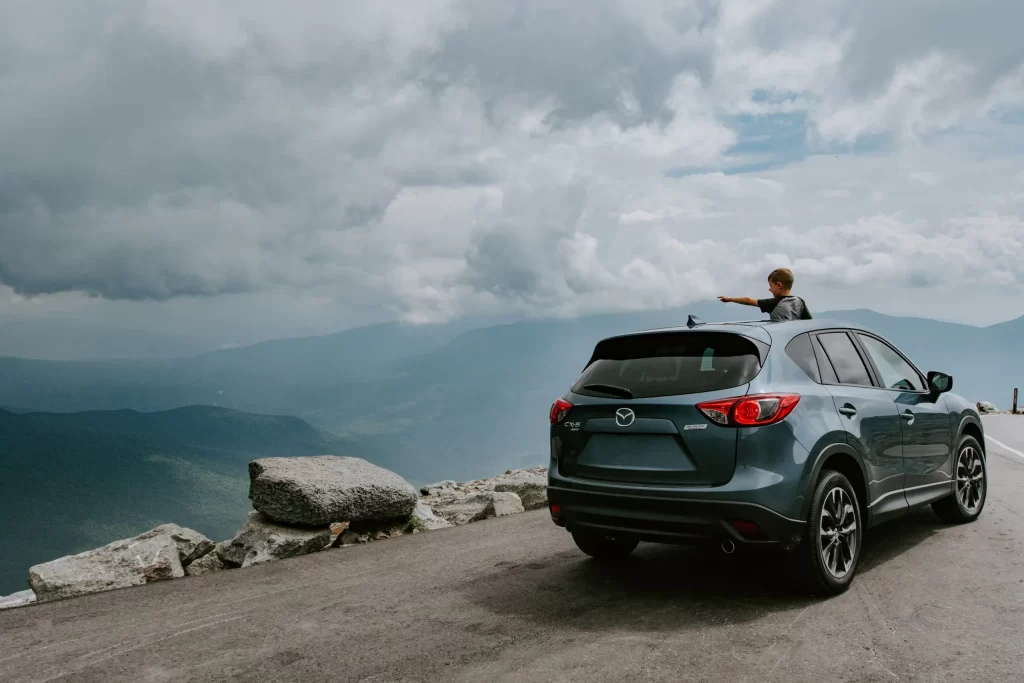 A child leans out of the sunroof of a parked gray Mazda SUV on a mountain road, pointing toward the misty, cloud-covered landscape and distant hills. Large rocks line the roadside.