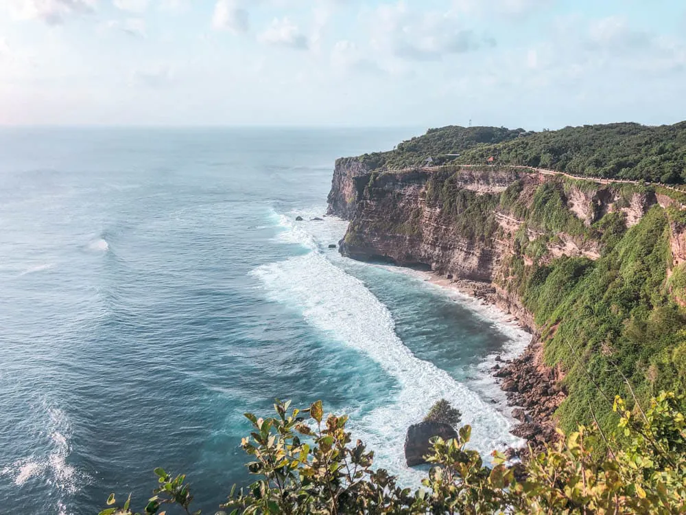 A scenic coastline with tall, green cliffs overlooking blue ocean waves crashing against the rocks, under a partly cloudy sky. Dense vegetation covers the top of the cliffs in the distance.