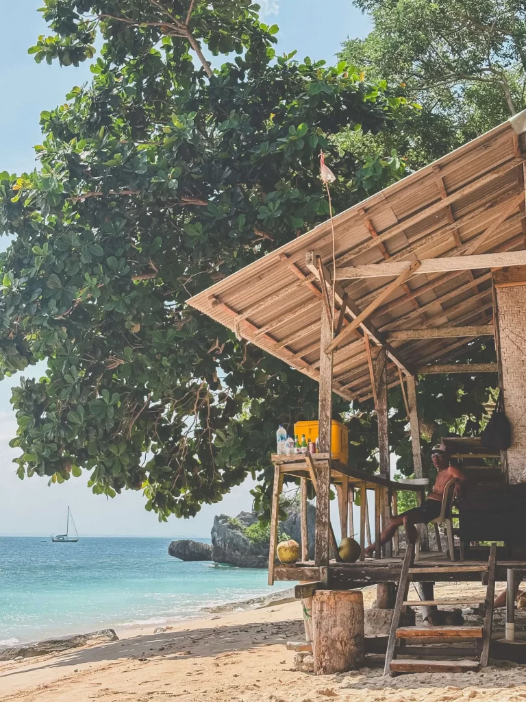 A rustic wooden hut stands on a sandy beach beside turquoise water, shaded by a large tree. A person sits on the porch, and a sailboat floats in the distance under a clear, sunny sky—evoking the laid-back charm of Thomas Beach Uluwatu.