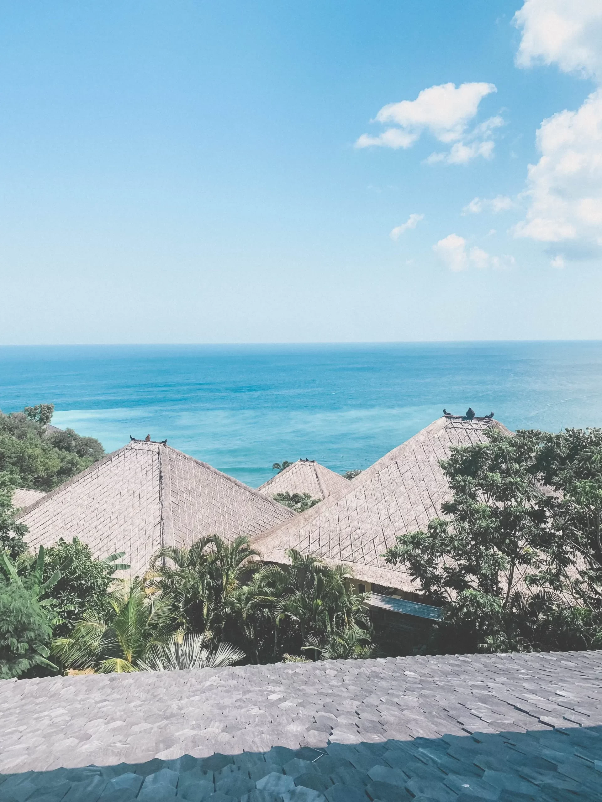 Thatched rooftops surrounded by green trees overlook a calm blue ocean under a partly cloudy sky, offering classic Mana Views Uluwatu charm in this serene surf villa.