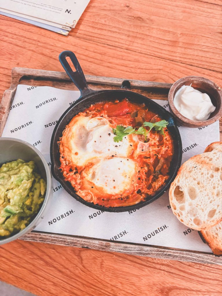 A skillet with baked eggs in tomato sauce, served with sliced bread, a bowl of guacamole, and a side of sour cream on a wooden tray—just one of the wholesome dishes you can enjoy at Nourish Cafe Uluwatu.