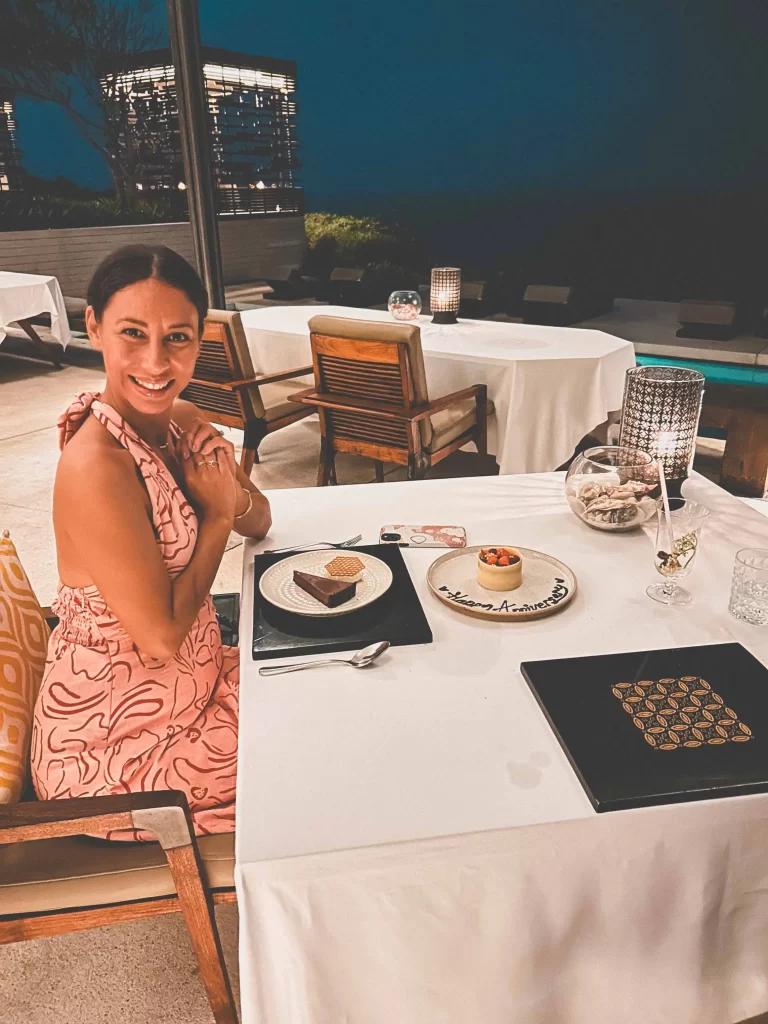 A woman in a pink dress smiles at a restaurant table with desserts, menus, and a drink at CIRE Restaurant Uluwatu, with outdoor seating and modern lighting in the background at night.