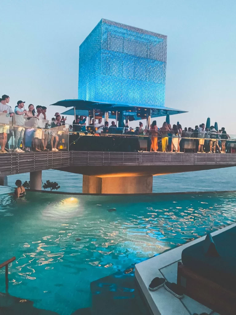 A lively crowd gathers on a modern, elevated bar with blue lights at Savaya Uluwatu, overlooking a swimming pool and the ocean at sunset.