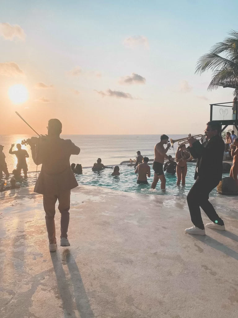 Two musicians play string instruments by the pool at sunset at El Kabron, while people swim and relax. Palm trees and the ocean are visible in the background, creating a lively, tropical atmosphere.