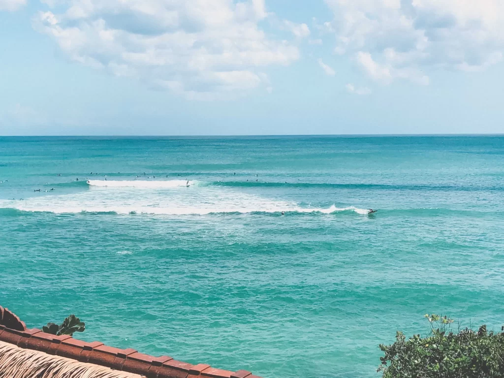 View of turquoise ocean waves with surfers in the distance at Uluwatu Beach, under a partly cloudy sky. Some greenery and a roof edge appear in the foreground.