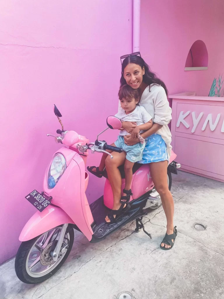 A woman and a young child sit on a pink scooter in front of a pastel pink wallβperhaps outside Kynd Cafe. Smiling at the camera, they both look happy and relaxed, capturing a perfect moment for any Bali itinerary.