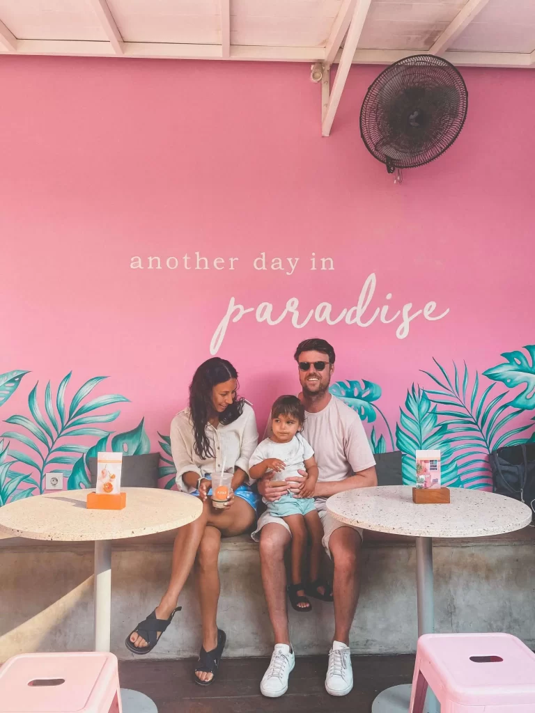 A smiling family of three sits at Kynd Cafe Bali, a must-stop on any Bali itinerary, with a pink wall that says another day in paradise and bold tropical leaf designs. They enjoy drinks together, looking happy and relaxed.